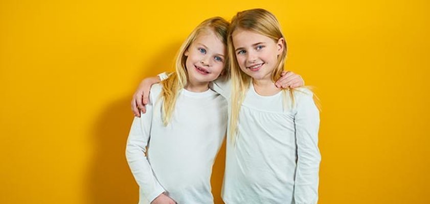 Zwei blonde Mädchen stehen Arm in Arm vor einem gelben Hintergrund